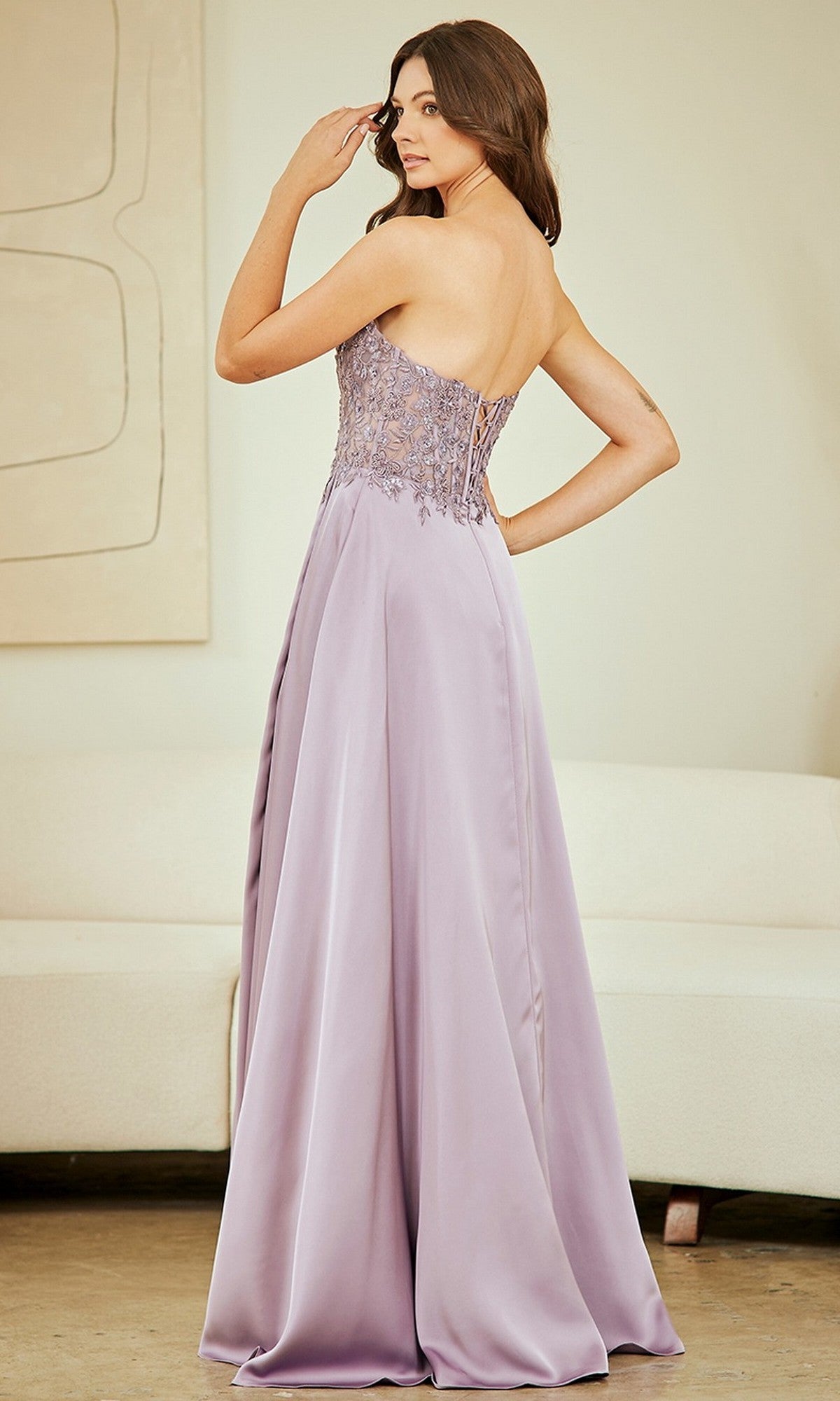 A woman, seen from behind indoors, wears the Amelia Couture 6204 long prom dress in lavender with a strapless beaded bodice and flowing skirt.